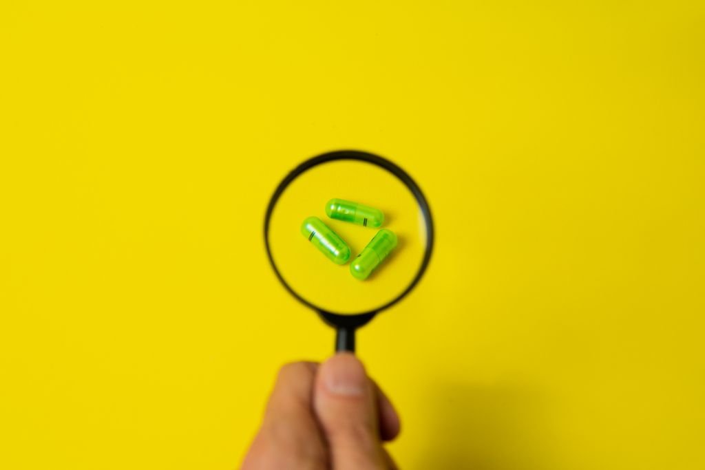 Top view of crop unrecognizable person holding magnifier over green pills placed on bright yellow surface in studio